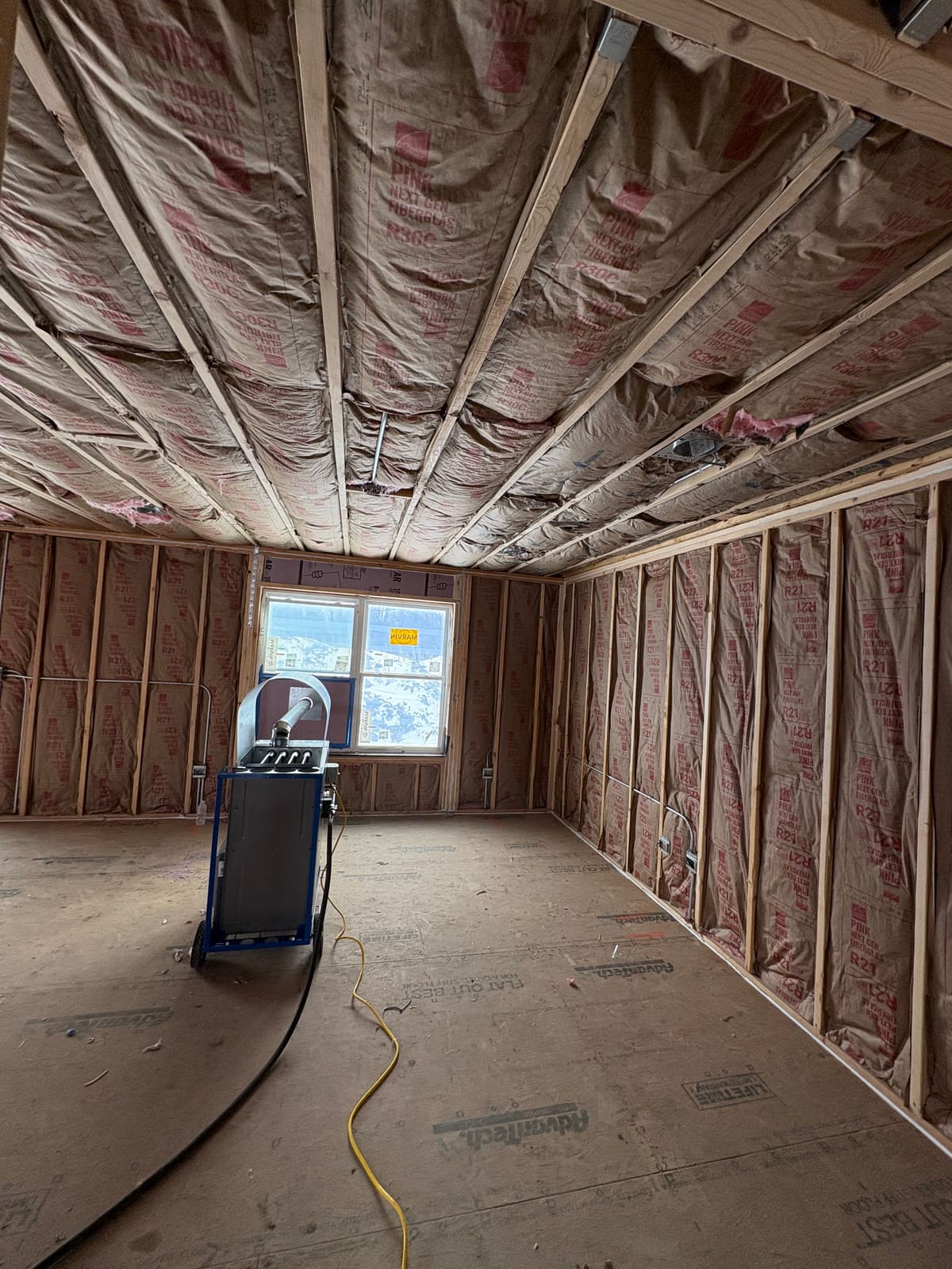 Kraft faced fiberglass batt insulation installed between the ceiling joists and in the wall stud cavities of a newly framed room near Elgin, IL, with snow outside the window and a plywood subfloor underfoot
