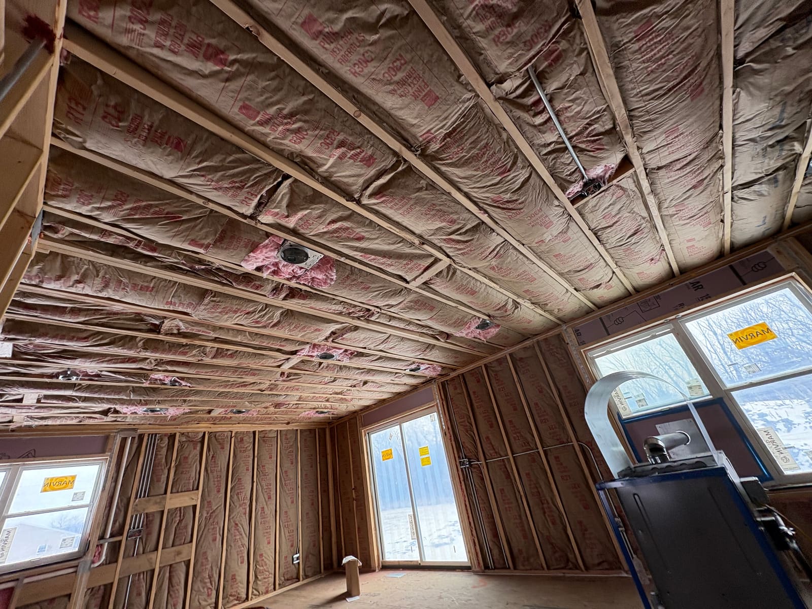 Kraft faced fiberglass batt insulation installed between the ceiling joists and the wall studs of a newly framed room, with snow on the ground outside the windows