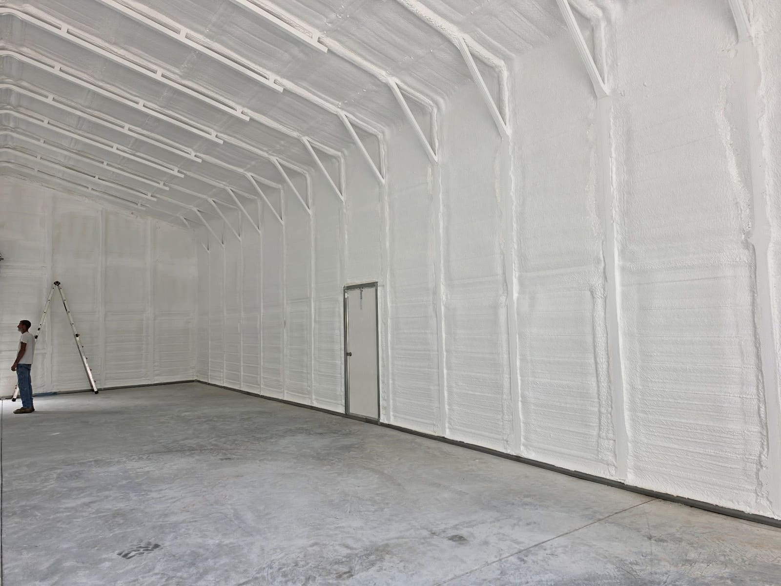 Interior of an empty pole barn with white spray foam insulation covering the sidewalls, the roof line and the steel framing, and a worker standing beside an extension ladder at the far end