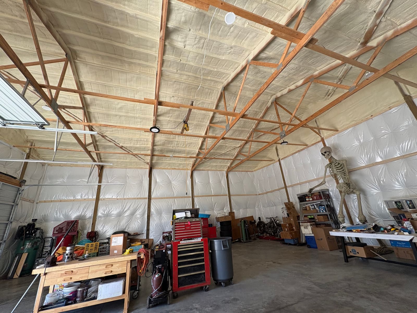 Wide interior view of an insulated pole barn workshop, with spray foam across the roof deck, white faced insulation panels on the walls and an overhead garage door on the left