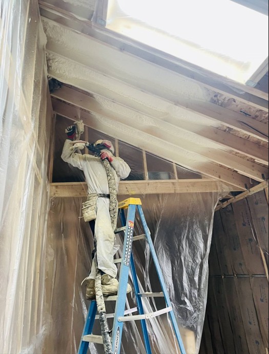 Solar Spray Foam Insulation technician in a protective suit spraying foam insulation onto a sloped ceiling from a step ladder in Kane County, IL, with plastic sheeting protecting the surrounding room