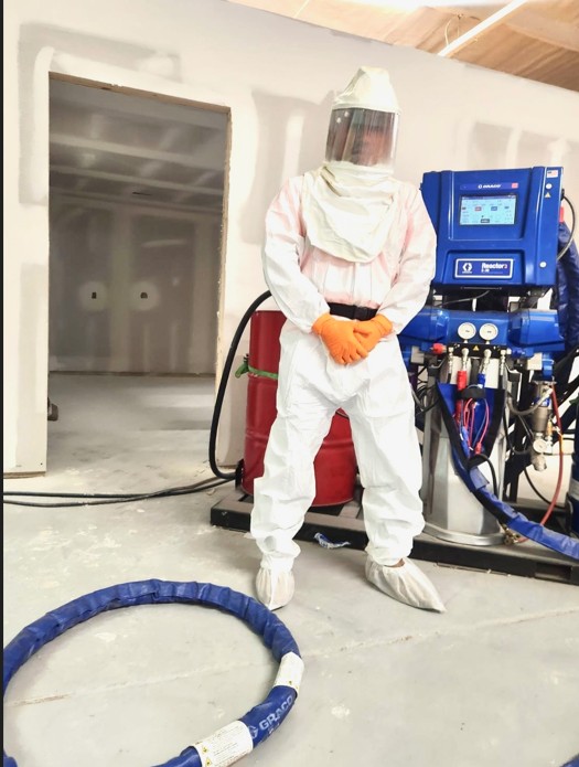 Solar Spray Foam Insulation technician in a full hooded protective suit and face shield standing beside the spray foam rig and heated hose set up inside a drywalled interior in Kane County, IL
